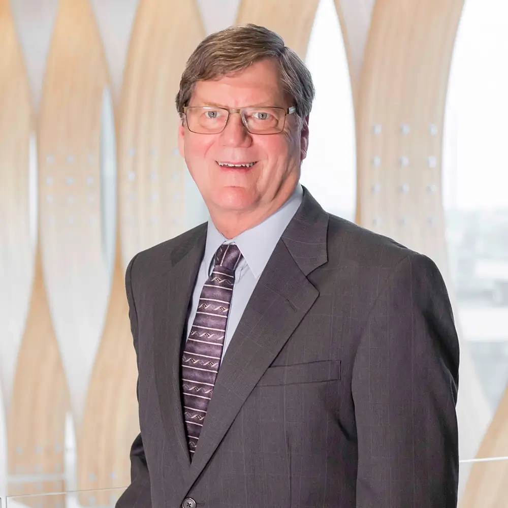 A middle-aged man with short brown hair and glasses, Tim Meyer, wearing a gray suit, patterned tie, and light blue shirt, smiles while standing in front of a modern background with curved wood panels and large windows—an OD professional at work.