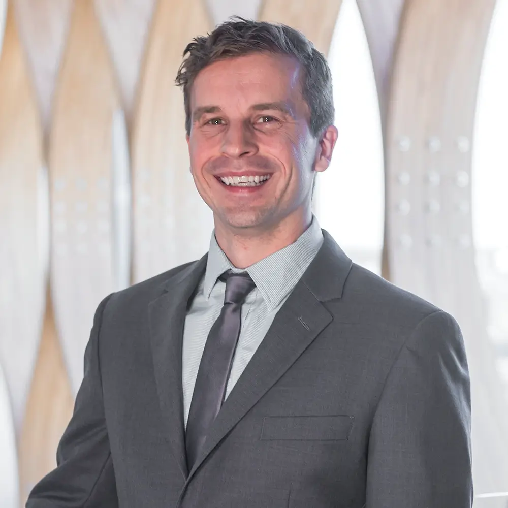 A man in a gray suit and tie smiles at the camera, standing in front of a modern architectural backdrop with curved wooden panels and bright lighting—Dr. Caleb Nabower, DPT, exudes confidence and professionalism.