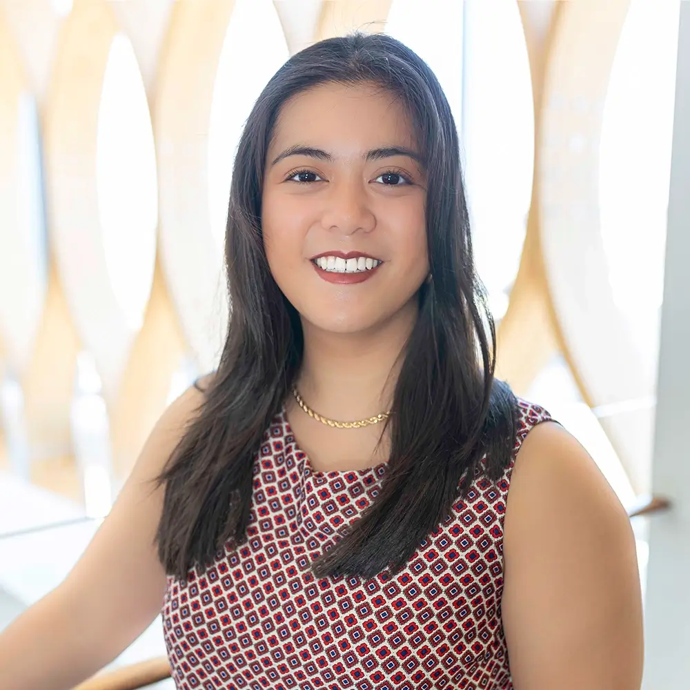 Henamari Ybay, DPT, with long dark hair, wears a patterned sleeveless top and a gold necklace as she smiles at the camera in a bright modern space with wooden and white oval designs in the background.