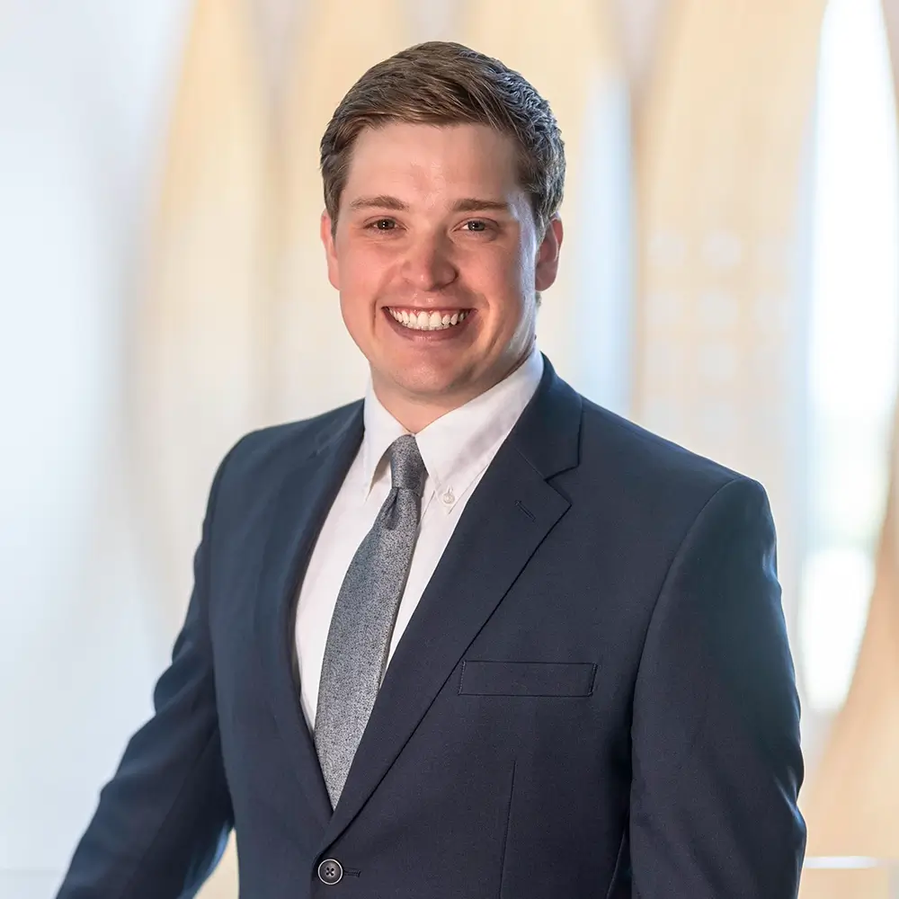 Jackson Buck, DPT, a young man in a dark suit, white shirt, and gray tie, smiles at the camera. He stands in a brightly lit, modern setting with blurred light-colored shapes in the background.