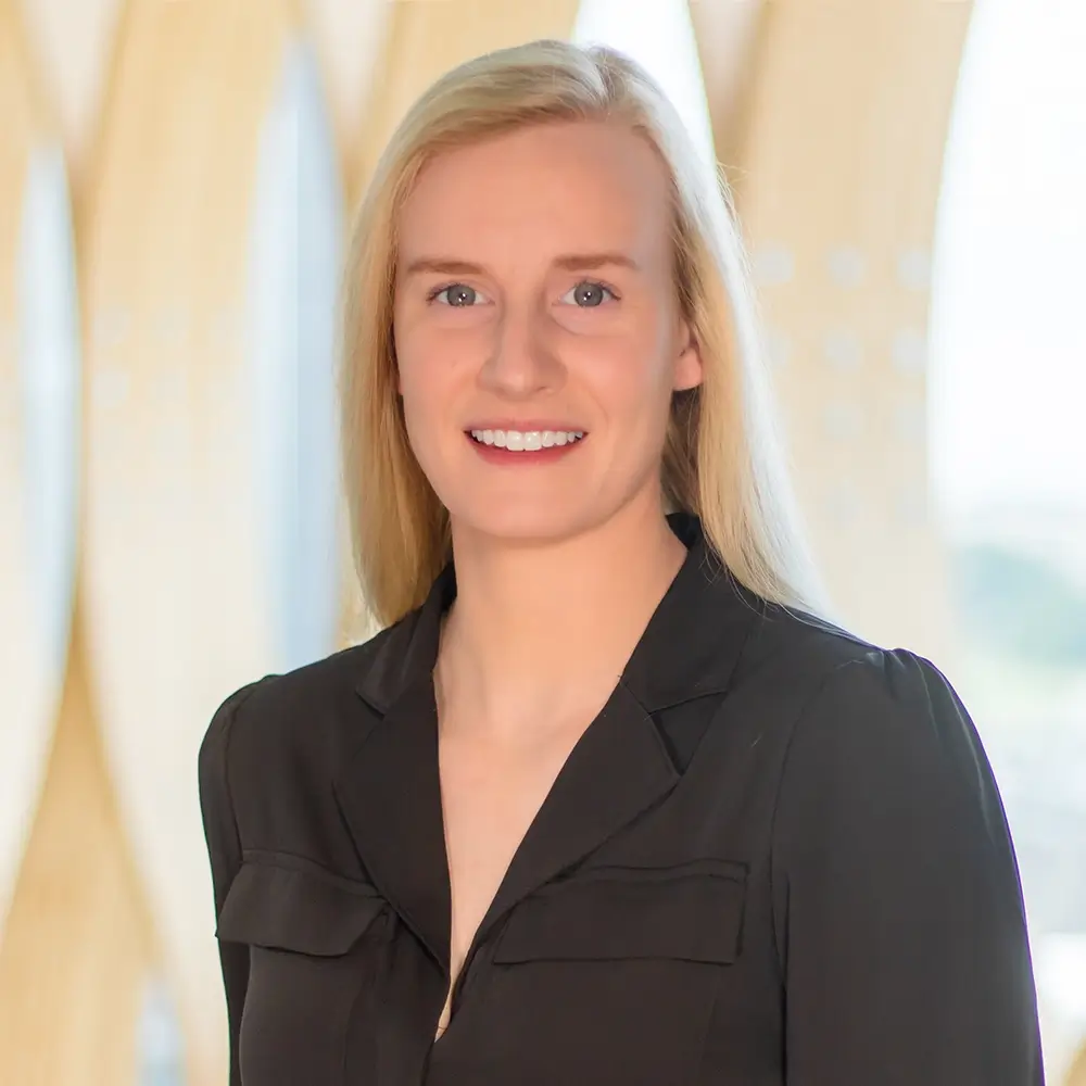 Mary Heimes, DPM, a woman with long blonde hair wearing a black blouse, stands indoors smiling against a softly blurred modern background with light wood and glass elements.