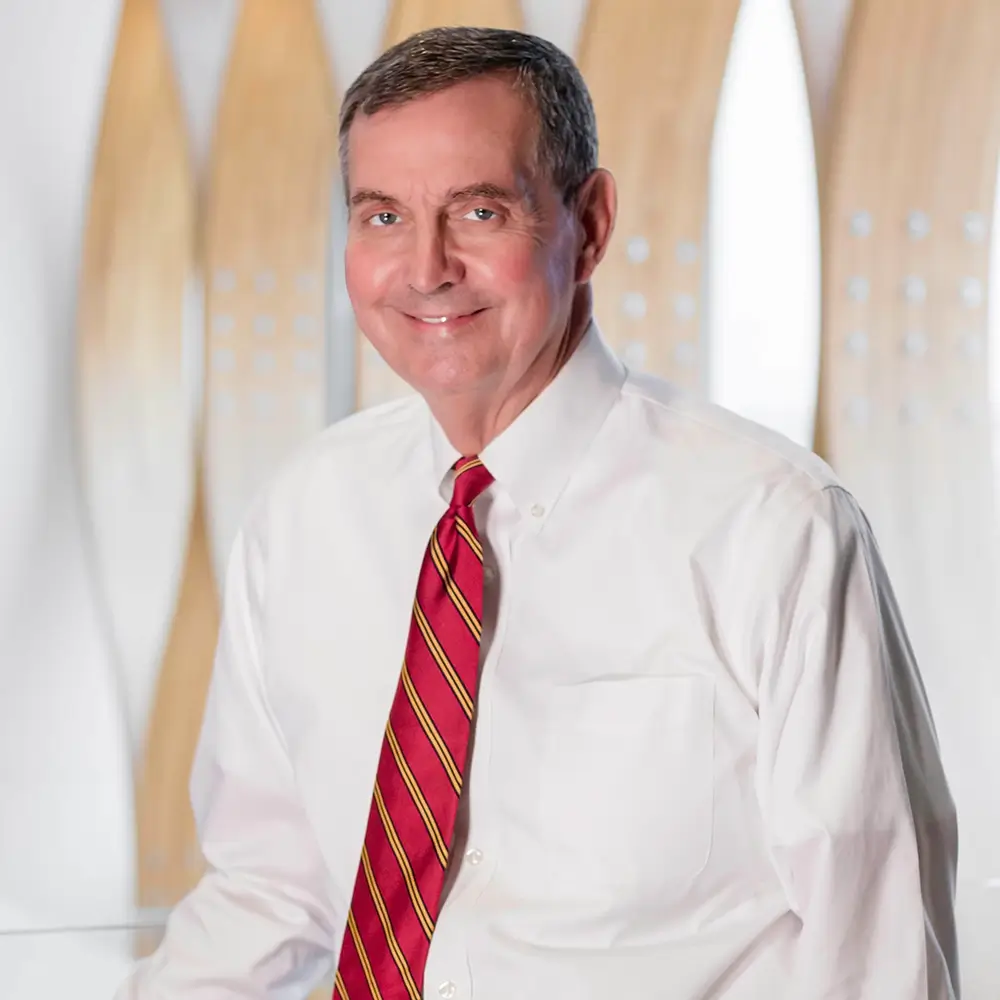 Dr. Richard Osterholm, MD, smiles at the camera in a white dress shirt and red striped tie, standing before a backdrop of vertical, light wood-patterned panels.