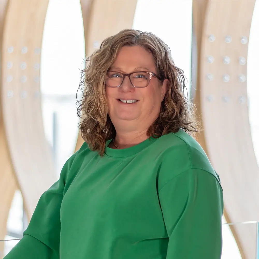 Risa Zimmerman, PA-C, with wavy, shoulder-length light brown hair and glasses, smiles at the camera in a bright green sweater. The modern wooden background and natural light add a warm touch to the scene.