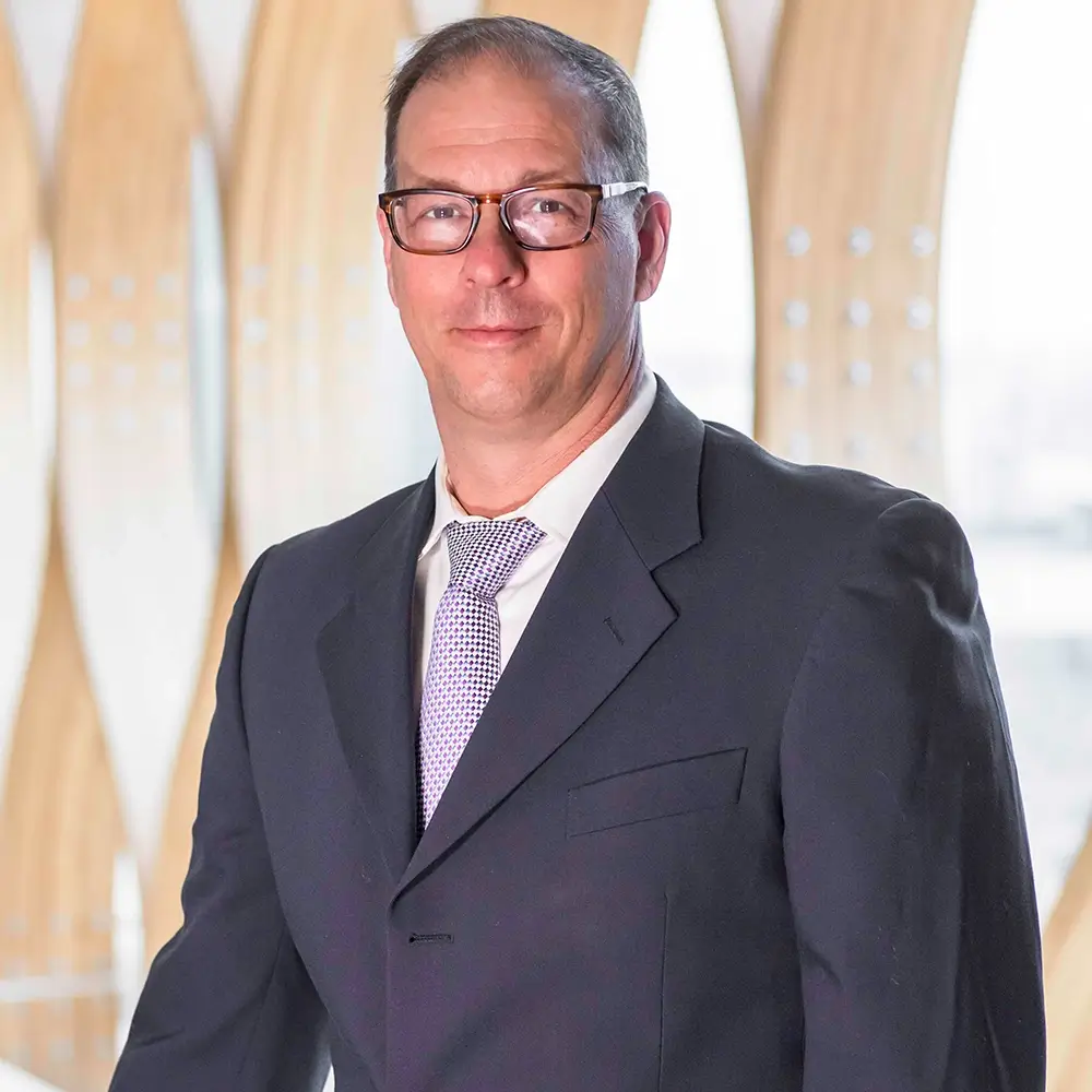Dr. Robert Recker, MD, wearing glasses, a dark suit, white shirt, and patterned tie, stands indoors before a modern light wood architectural background, smiling slightly at the camera.