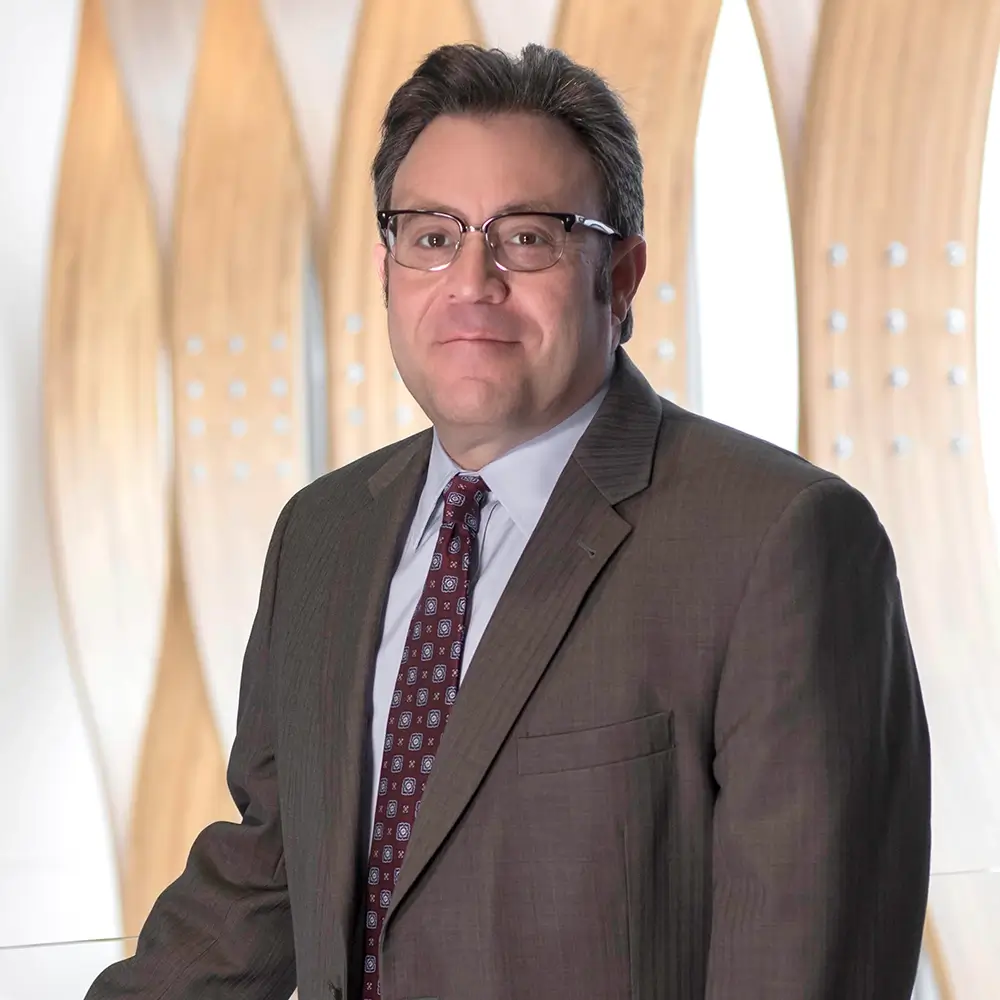 Dr. Steven Zuber, MD, wearing glasses and a brown suit with a patterned tie, stands in front of a modern, light-colored wooden wall with curved design elements. He is looking at the camera and smiling slightly.