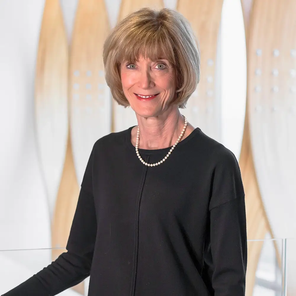 Dr. Susan Scholer, MD, with short light brown hair, wearing a black top and pearl necklace, stands smiling in front of a modern, light-colored wooden architectural background.