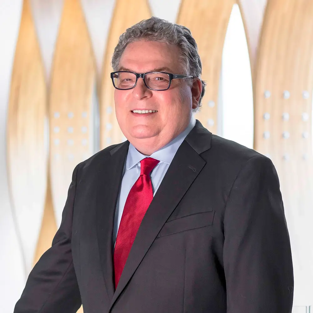 Dr. Timothy Crnkovich, MD, wearing glasses, a dark suit, light blue shirt, and red tie, stands smiling in front of a modern background with light wood and white accents.