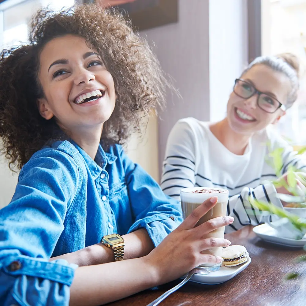 Two young women smiling at a cafe table; the one in the foreground holds a latte with a macaron on her saucer, wearing a denim shirt and a gold watch.