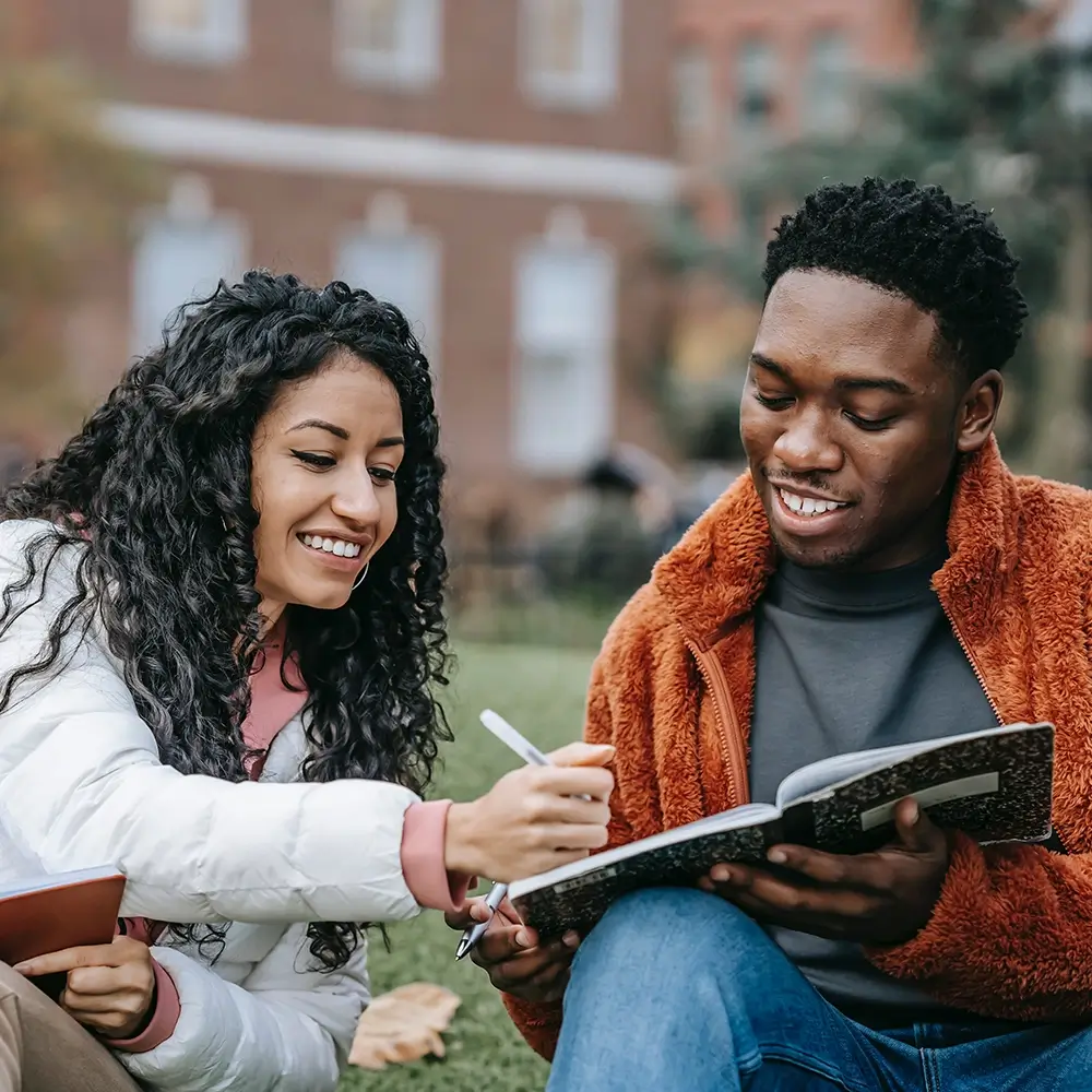 Two students sit on grass outside, studying together and smiling as one writes in a notebook while the other reads.