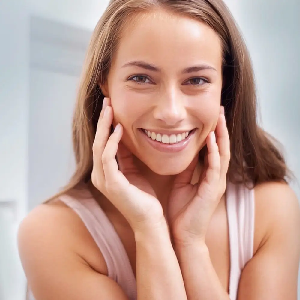 A woman with long brown hair smiles brightly, gently touching her face with both hands. She is wearing a light-colored sleeveless top and is in a softly lit, indoor setting.