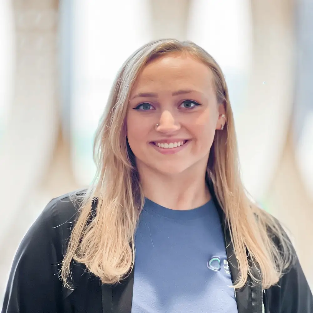 A young woman with long blonde hair smiles at the camera. She is wearing a dark jacket over a blue shirt and stands in front of a softly blurred, light-colored background.