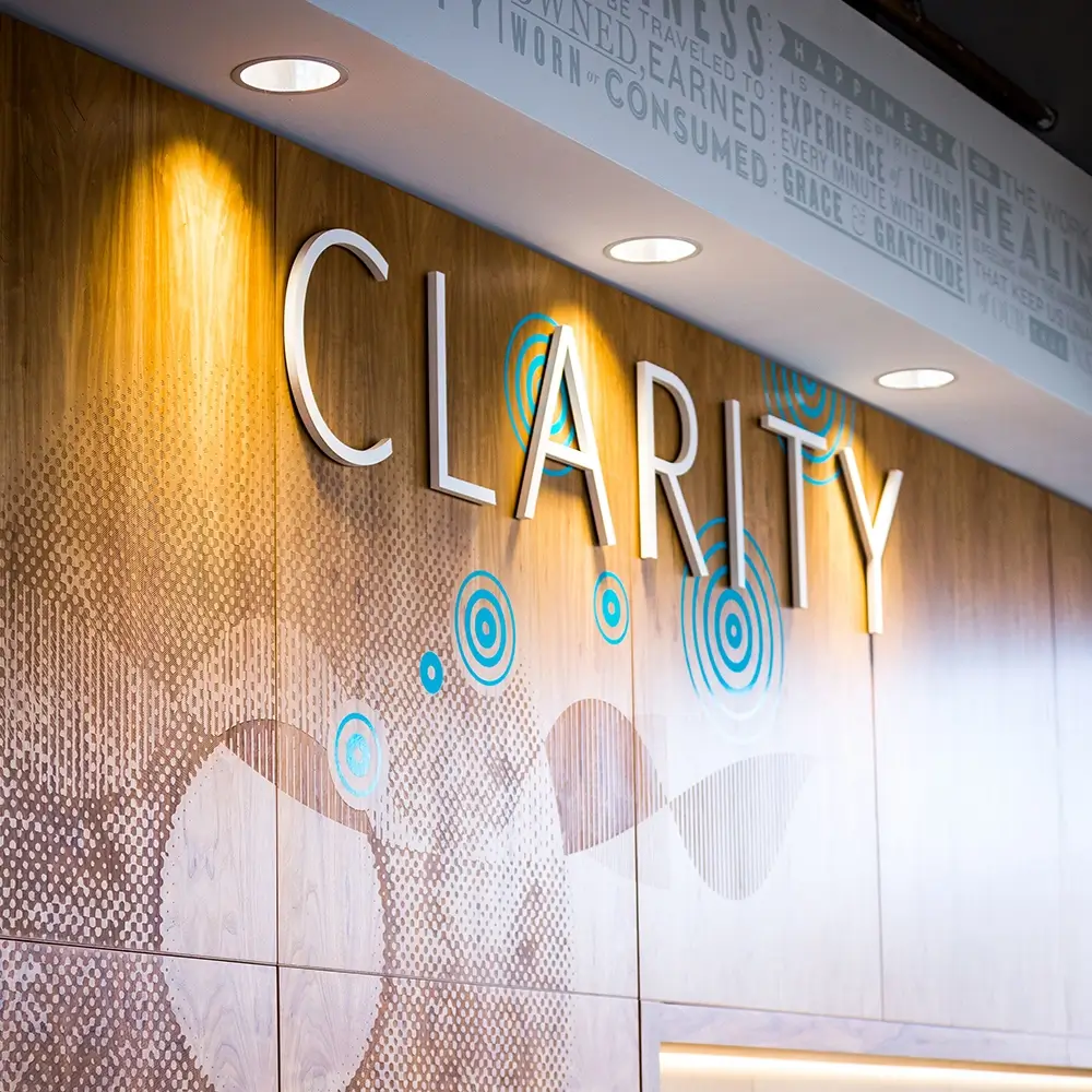 Wood-paneled wall with the word CLARITY in large white letters, blue and beige abstract patterns, and spotlights shining from the ceiling above. Upper wall features motivational words in smaller text.