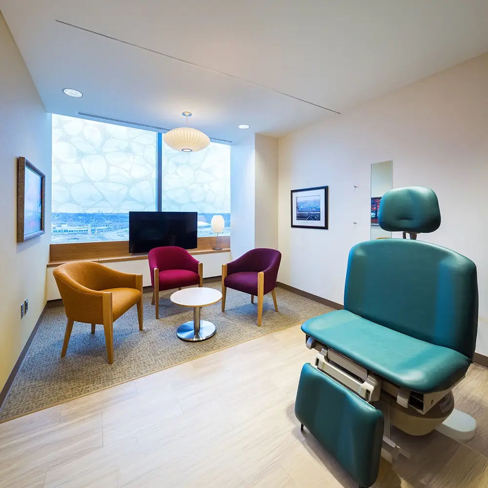 A modern medical exam room with a teal exam chair, three colorful chairs around a small table, large window, wall art, and a TV. The space is clean, bright, and welcoming.