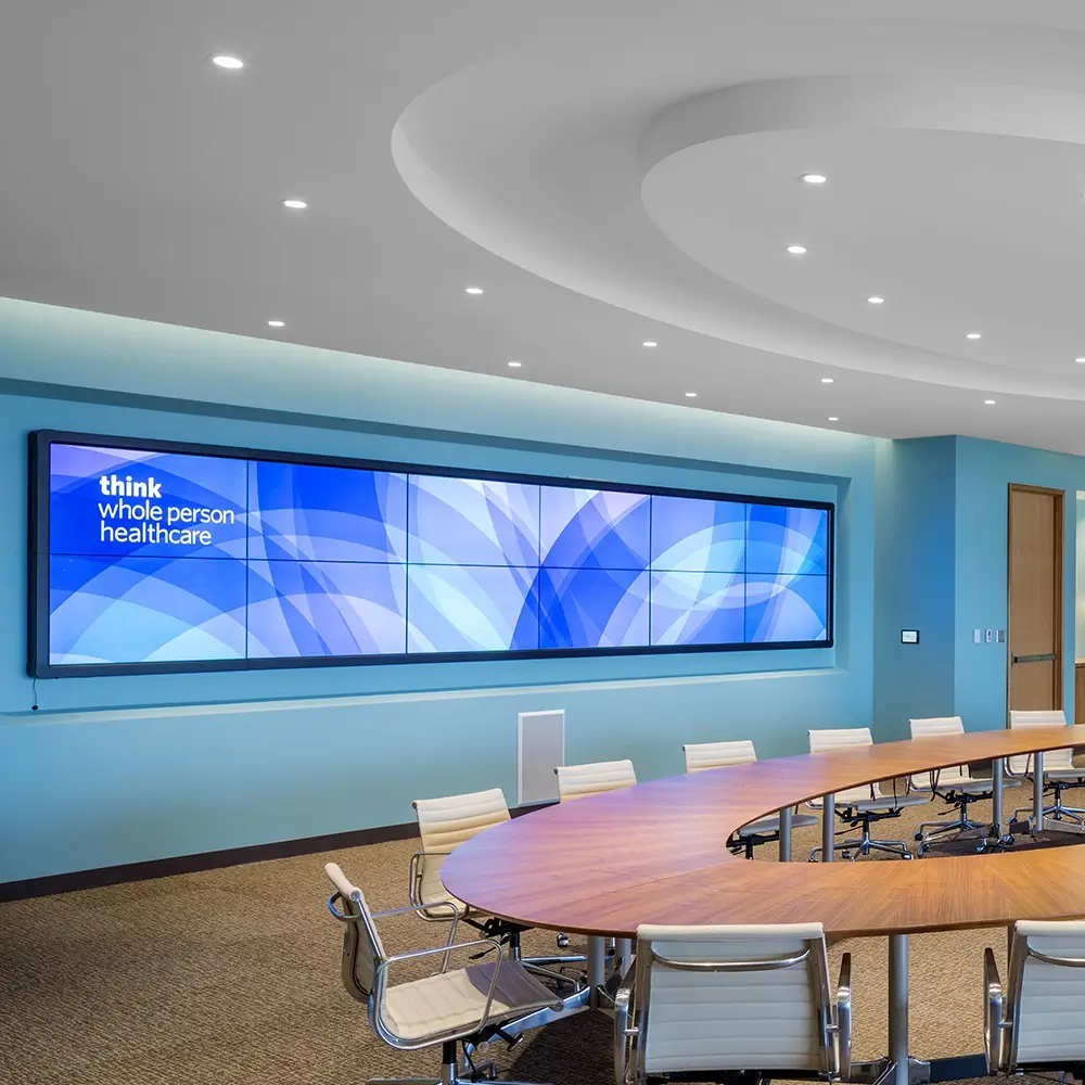 A modern conference room with a large oval table, beige chairs, and a wide digital screen on the wall displaying blue abstract graphics and the words think healthcare. The room features blue walls and recessed ceiling lights.