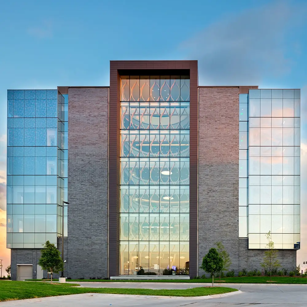 A modern building with a glass facade, symmetrical design, and brick accents. The entrance features large windows and patterns, with a clear sky and a few trees in the foreground.
