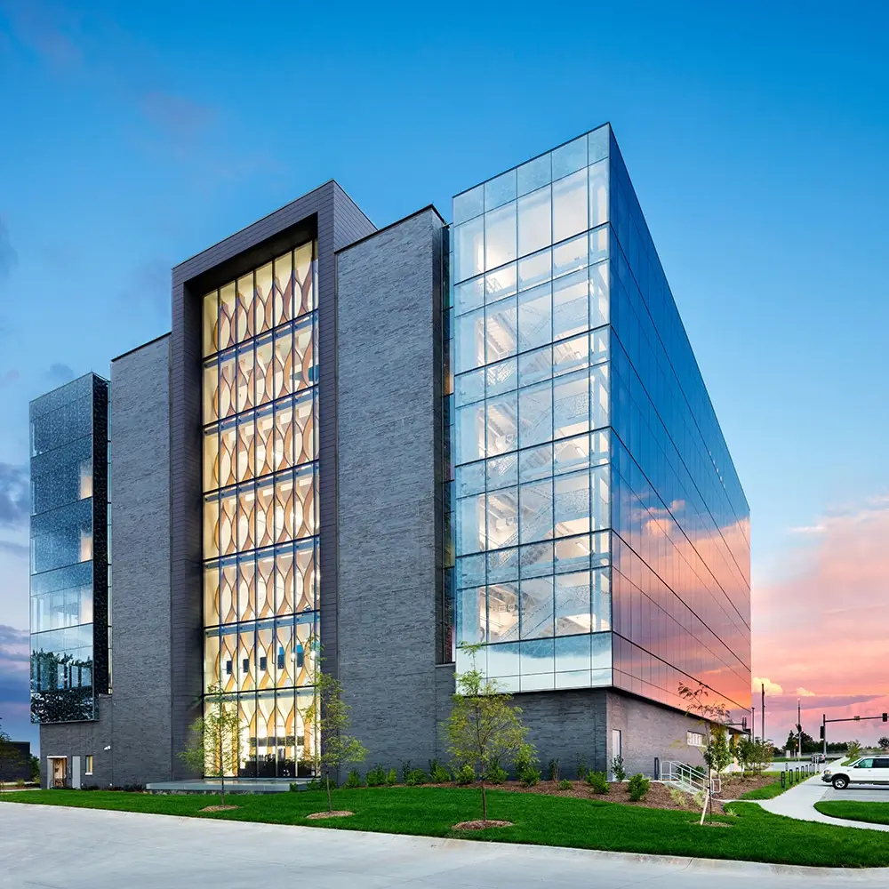 A modern glass and brick office building with large windows reflecting the sunset sky, ideal for think healthcare offices, surrounded by a neatly landscaped lawn and a paved driveway.