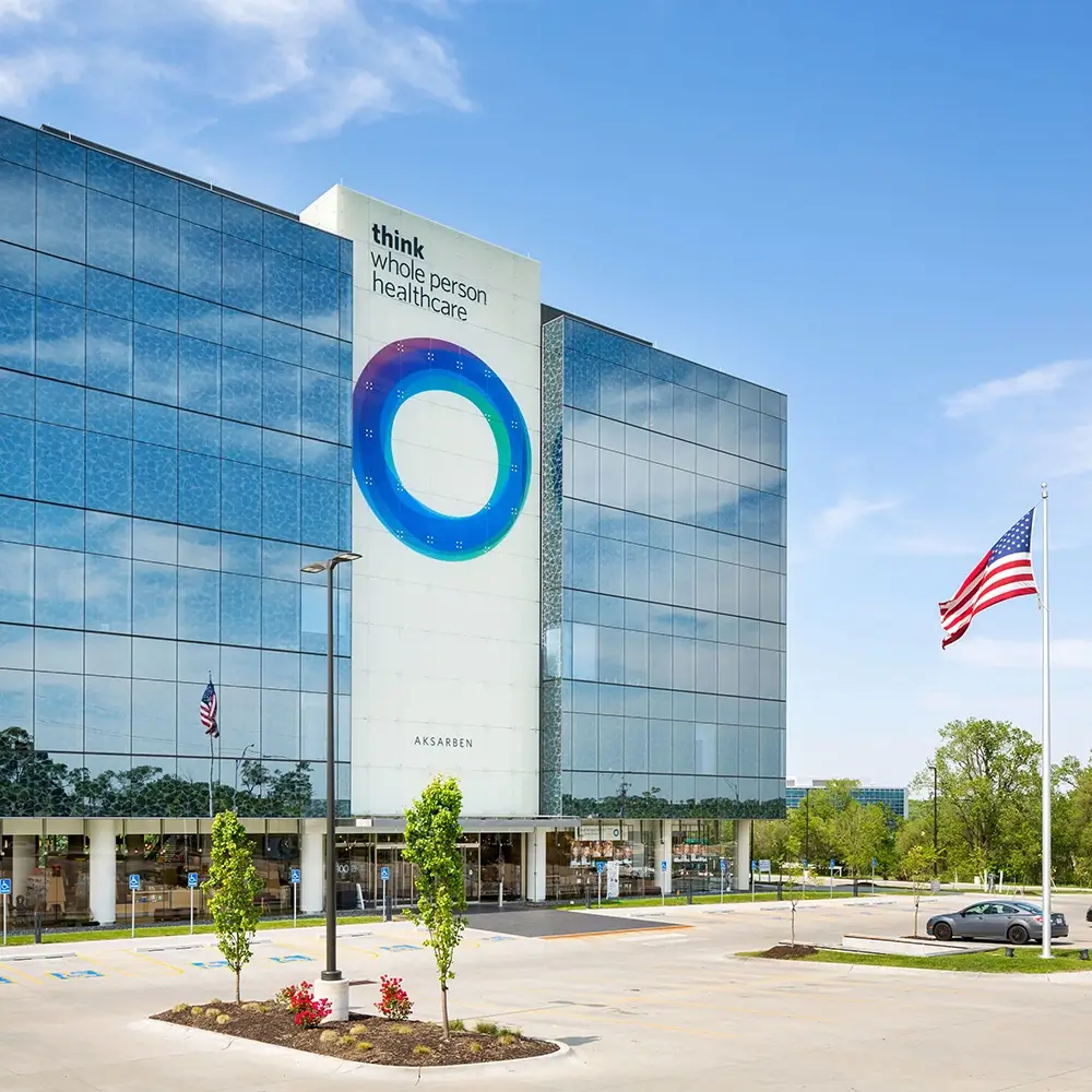 A modern glass building with think Whole Person Healthcare signage and a large circular design on the facade. An American flag is flying on a flagpole in the parking lot, with trees and clear blue sky in the background.