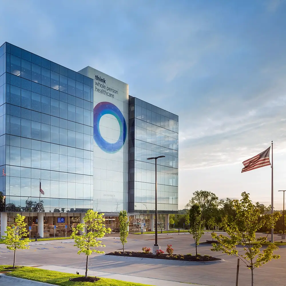 Modern glass building with a large circular logo and “think whole person healthcare” sign on the facade, surrounded by a parking lot, young trees, and an American flag on a flagpole; blue sky in the background.