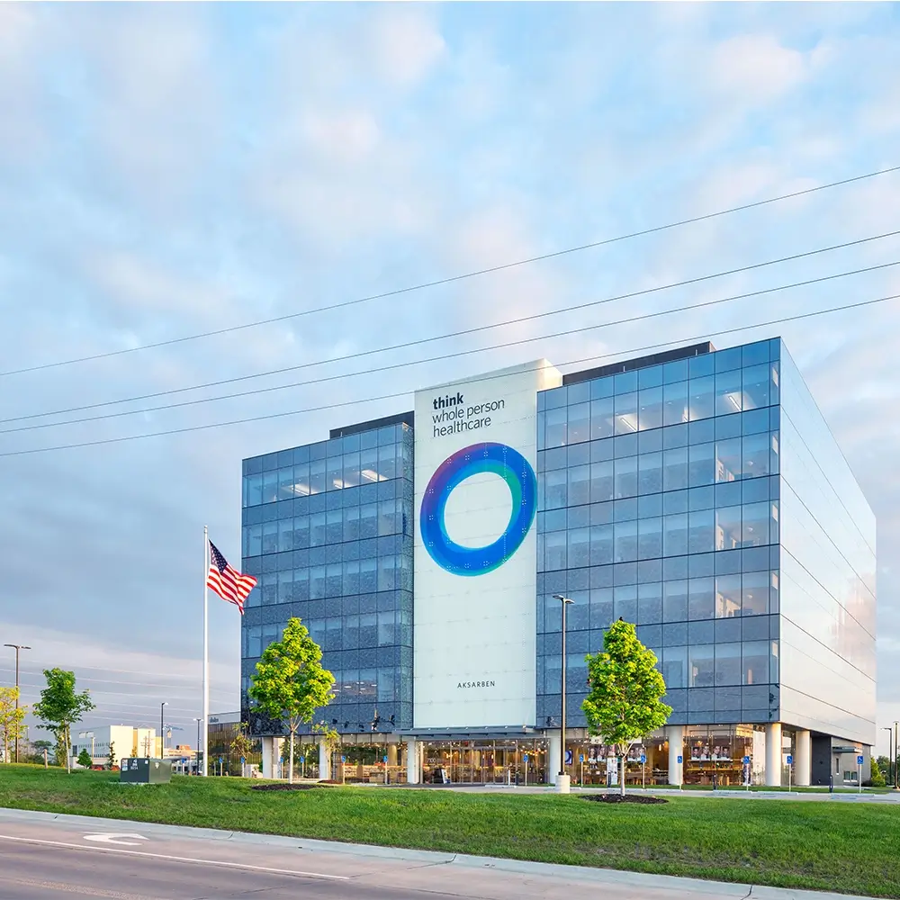 A modern glass office building with a circular logo and the words think whole person healthcare on the facade. An American flag is flying in front, and trees line the property along a road.