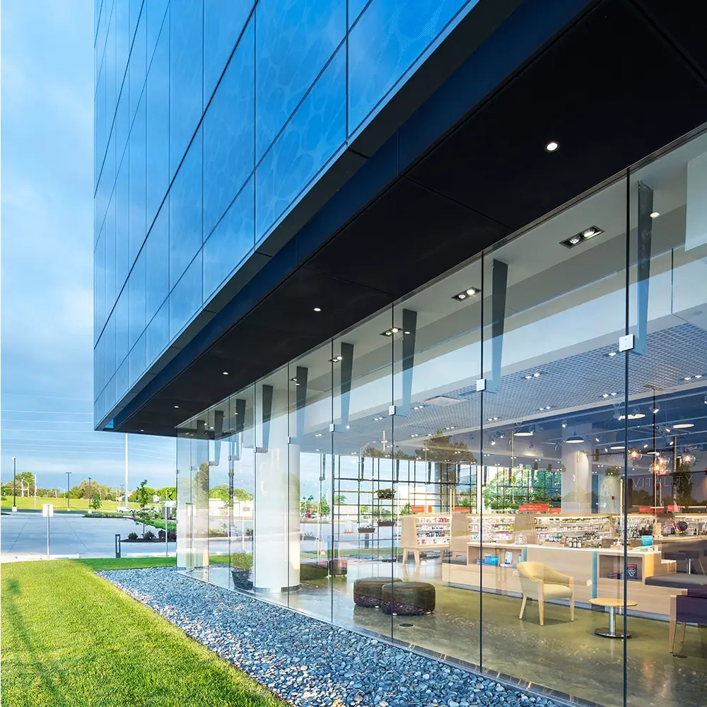 Modern glass building with floor-to-ceiling windows, showcasing a bright, contemporary interior with displays. Green grass and rocks line the exterior, reflecting the sky and surroundings on the glass facade.