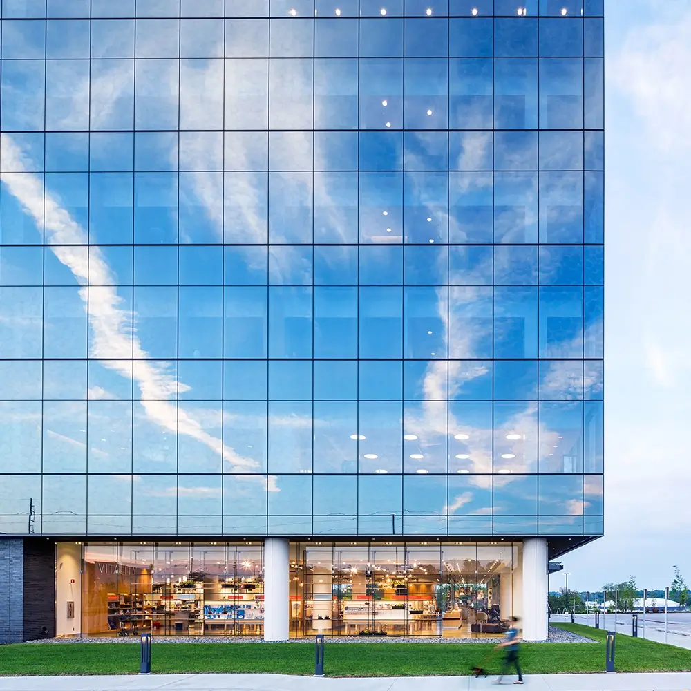 A modern glass building reflects a blue sky with white clouds. The lower floor has large windows revealing shelves and lights inside, while a person with a stroller passes by on the sidewalk outside.