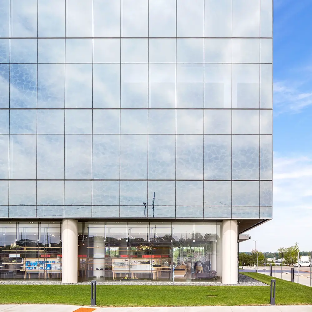 A modern glass office building with reflections of the sky, showing a ground-level café or office inside, green grass in front, and a blue sky with clouds above.