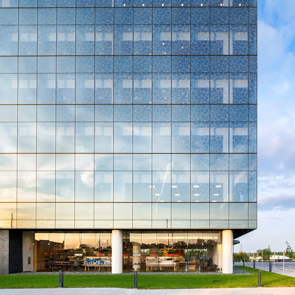 A modern glass office building reflects the sky and clouds. The ground floor features large windows revealing an interior designed for think healthcare, with desks, plants, and shelves. Two white columns support the upper structure.