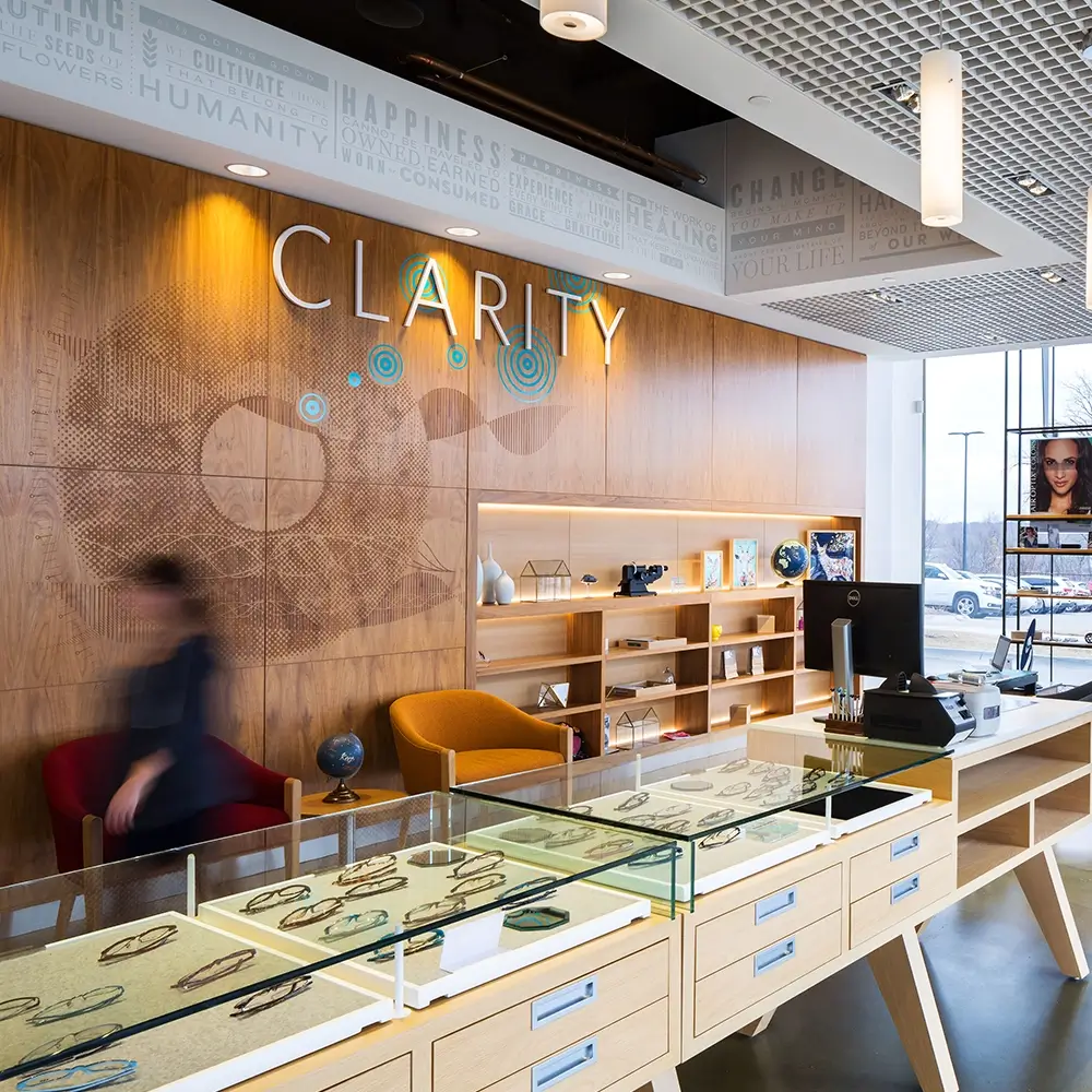 A modern optical store interior features wooden display cases with eyeglass frames, a wall with the word CLARITY, shelves with decorative items, and large windows letting in natural light. A person walks past in motion blur.