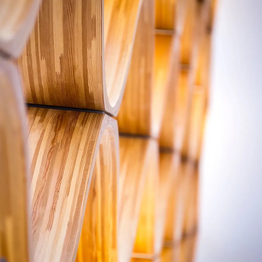 Close-up of curved wooden shelves forming a repeating geometric pattern, with warm lighting highlighting the wood grain and creating a modern, abstract design against a blurred light background.
