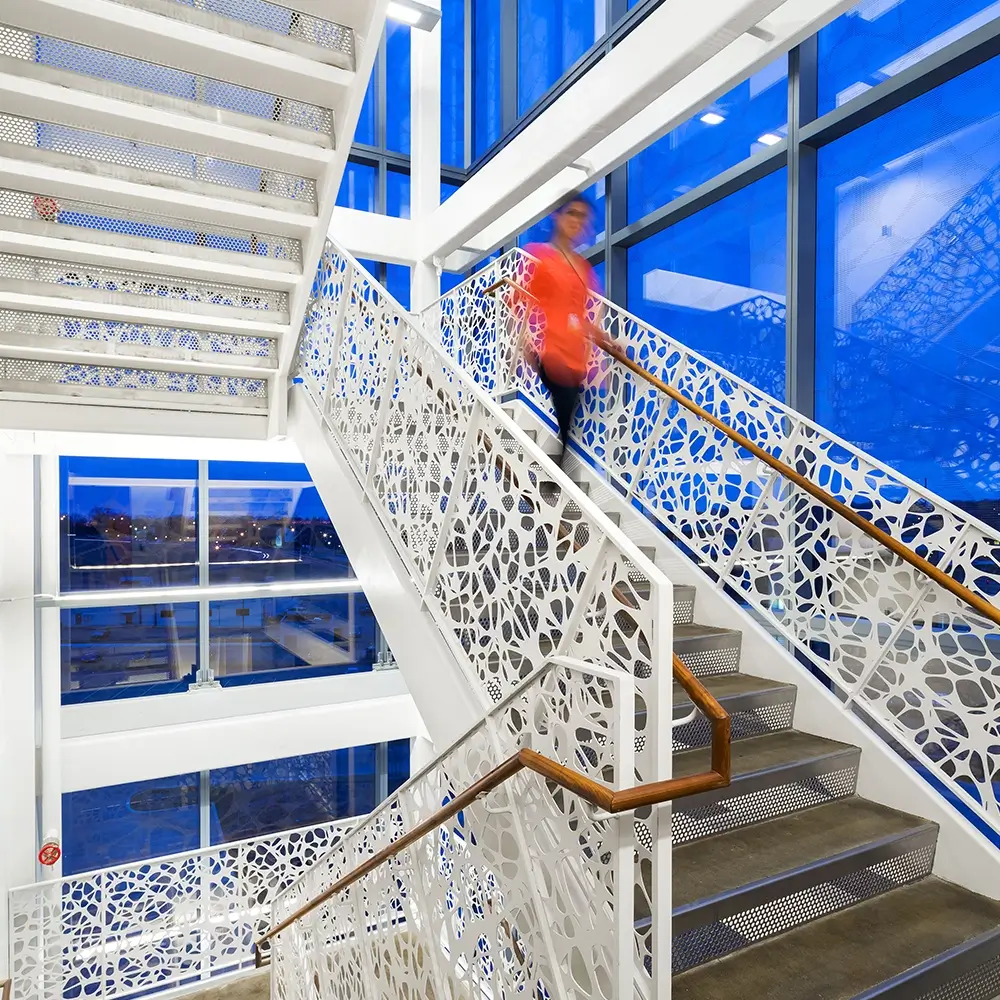 A person in a red top descends a modern staircase with intricate white geometric railings inside a bright think healthcare building, with large blue-tinted windows revealing a cityscape outside.