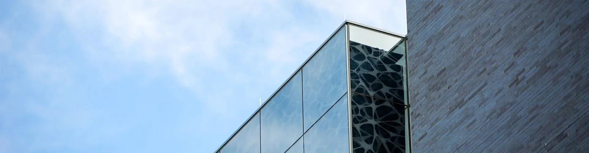 Modern building exterior with large glass windows reflecting patterned designs, next to a textured stone wall, against a blue sky with light clouds.