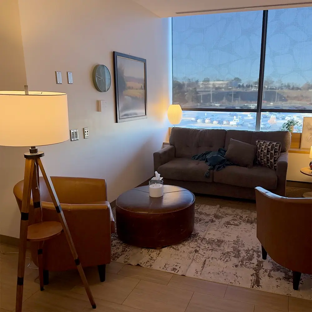 A cozy lounge with brown leather chairs, a gray sofa, a round ottoman, a standing lamp, and a large window letting in daylight. Throw pillows and a patterned rug add comfort to the warmly lit room.