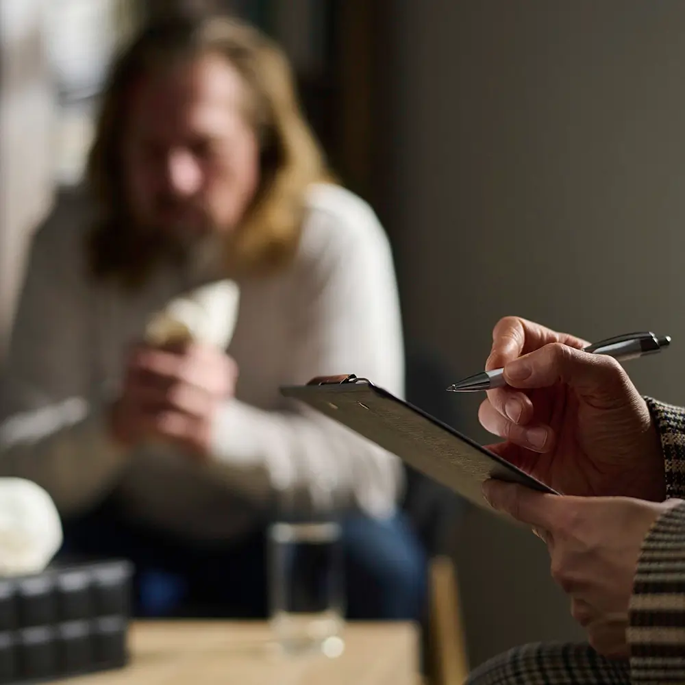 A person holds a clipboard and pen, taking notes, while another person sits in the background holding a tissue, suggesting a therapy or counseling session.