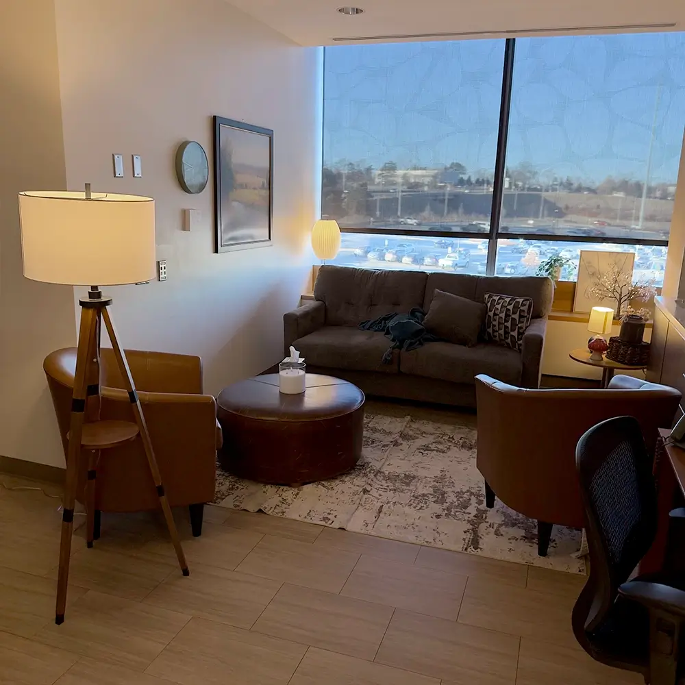 A cozy waiting room with brown chairs, a sofa, round coffee table, standing lamp, wall art, and large window letting in natural light. A cityscape and blue sky are visible outside.