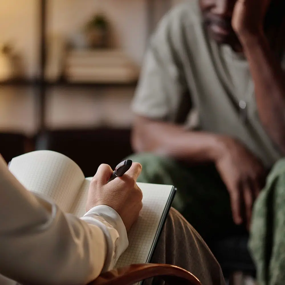 A person writes in a notebook while another person sits across from them, leaning forward with their hand on their face, suggesting a counseling or therapy session.