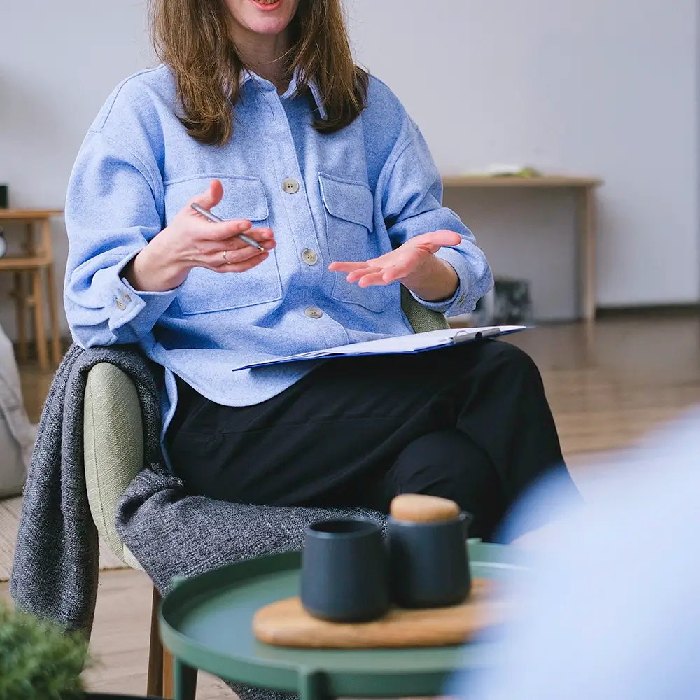 A person in a blue shirt sits in a chair holding a clipboard, gesturing with their hands while talking. A small table with a teapot and cups is in the foreground. The setting appears to be indoors and casual.