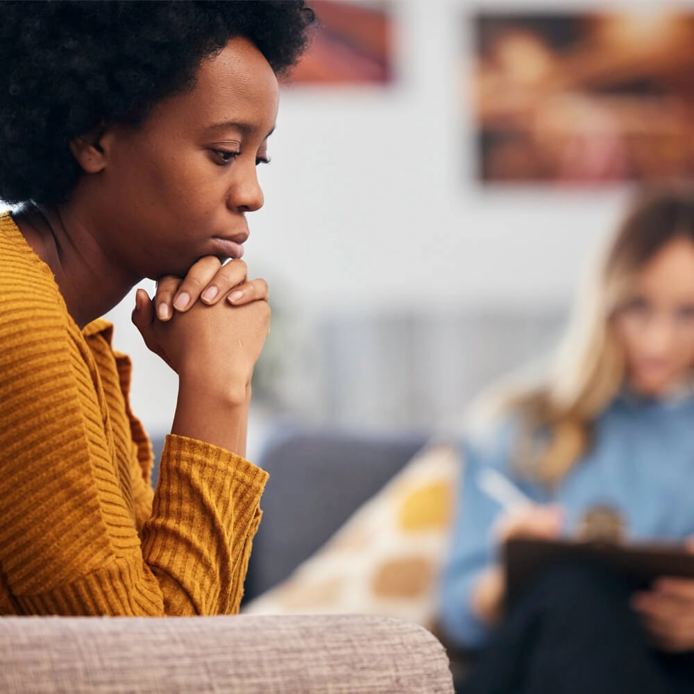 A woman in a yellow sweater sits on a couch, looking thoughtful and concerned with her hands clasped under her chin. In the blurred background, another person holds a clipboard, possibly a therapist.