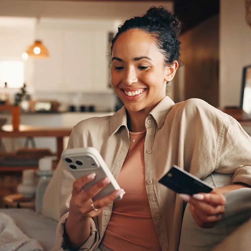 A smiling woman sits indoors holding a smartphone in one hand and a credit card in the other, appearing to make an online purchase in a cozy, well-lit living room.