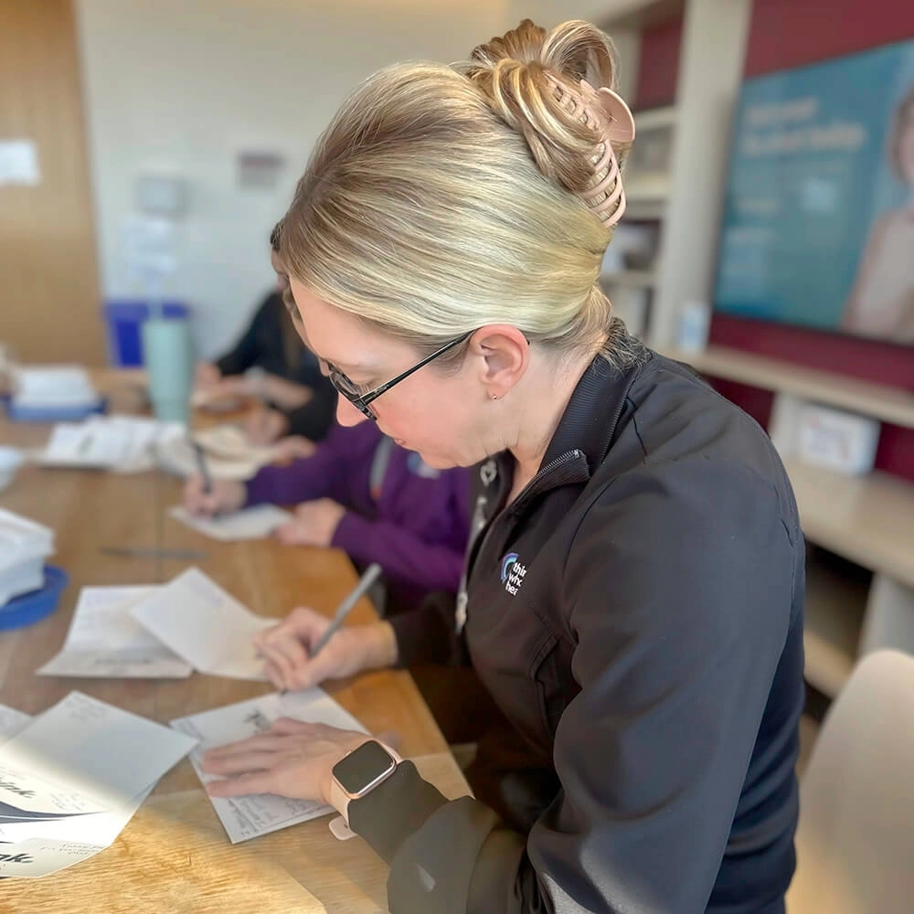A woman with blonde hair tied up, wearing glasses and a black jacket, writes on papers at a table. Other people are also working in the background. The setting appears to be an office or classroom.