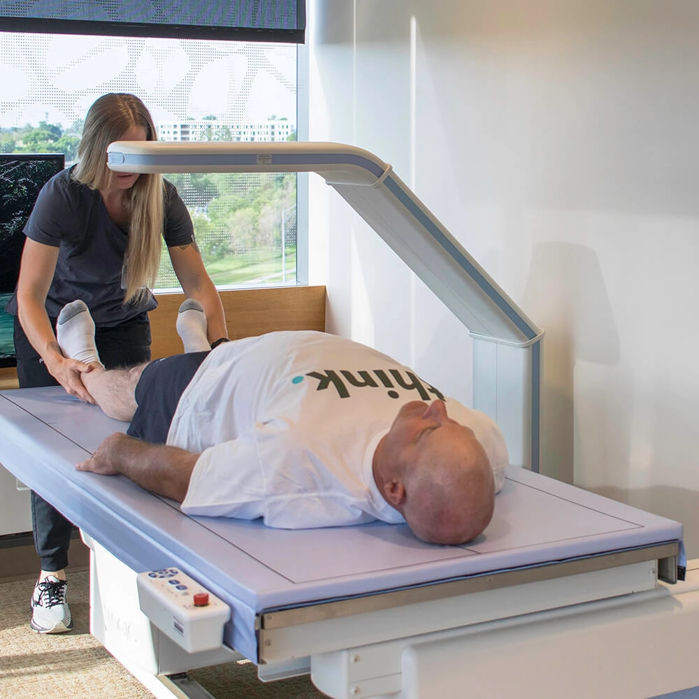 A man lies on his back on a medical scanning table while a healthcare professional adjusts his legs. The man wears a white T-shirt, and the room has large windows with a view of trees outside.