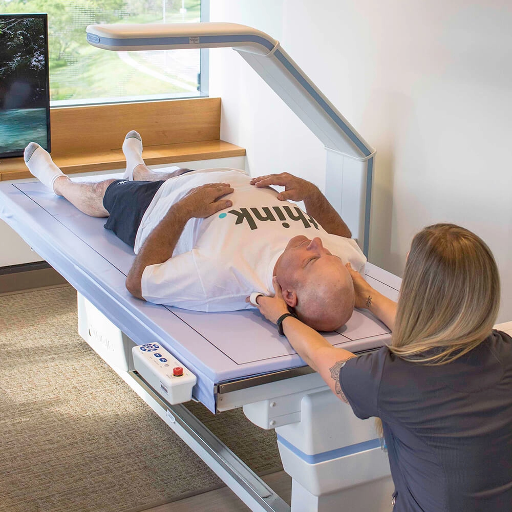 A person lies on their back on a medical imaging table while a healthcare professional adjusts their head position, preparing them for a scan in a bright room with large windows.