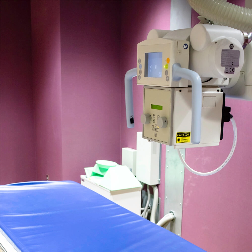 A medical X-ray machine with control panel and handles is mounted on a wall above a blue examination table in a room with pink walls. Nearby are medical supplies and equipment.