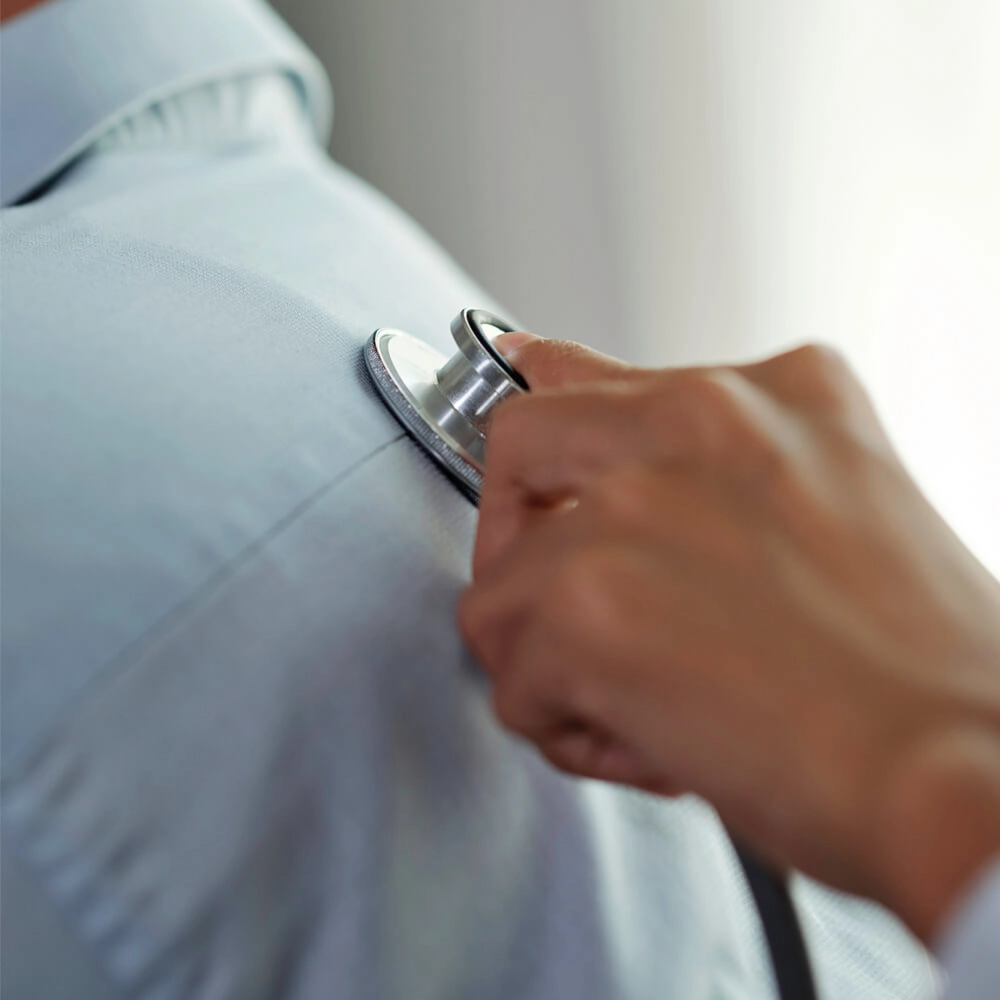 A close-up of a hand holding a stethoscope to the chest of a person wearing a light blue shirt, suggesting a medical examination or check-up.
