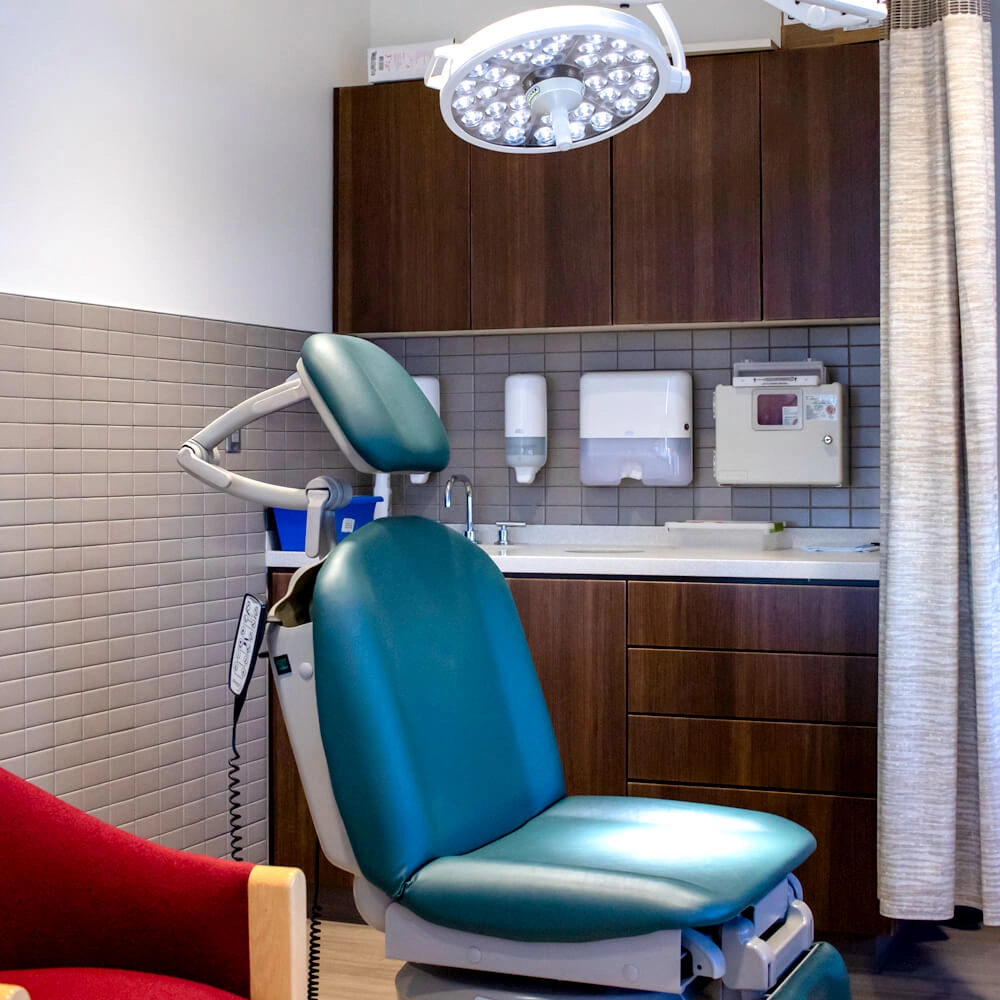A modern dental or medical exam room with a teal reclining chair, overhead exam light, wooden cabinets, sink, soap dispensers, and medical equipment against a tiled wall.