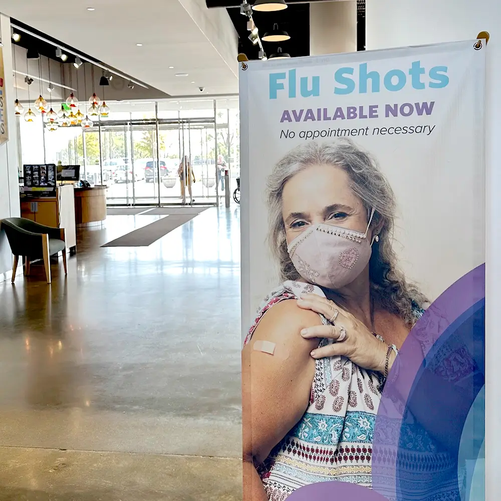 A banner in a lobby displays a woman wearing a mask and showing her arm with a bandage, promoting flu shots with the text: Flu Shots Available Now. No appointment necessary.