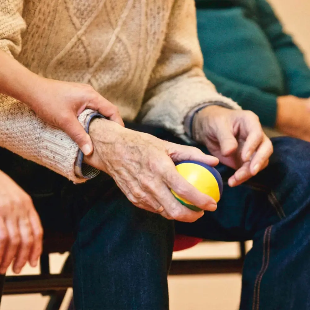 A person gently holding the arm of an elderly individual, who is seated and holding a small yellow and blue ball, suggesting support and care.