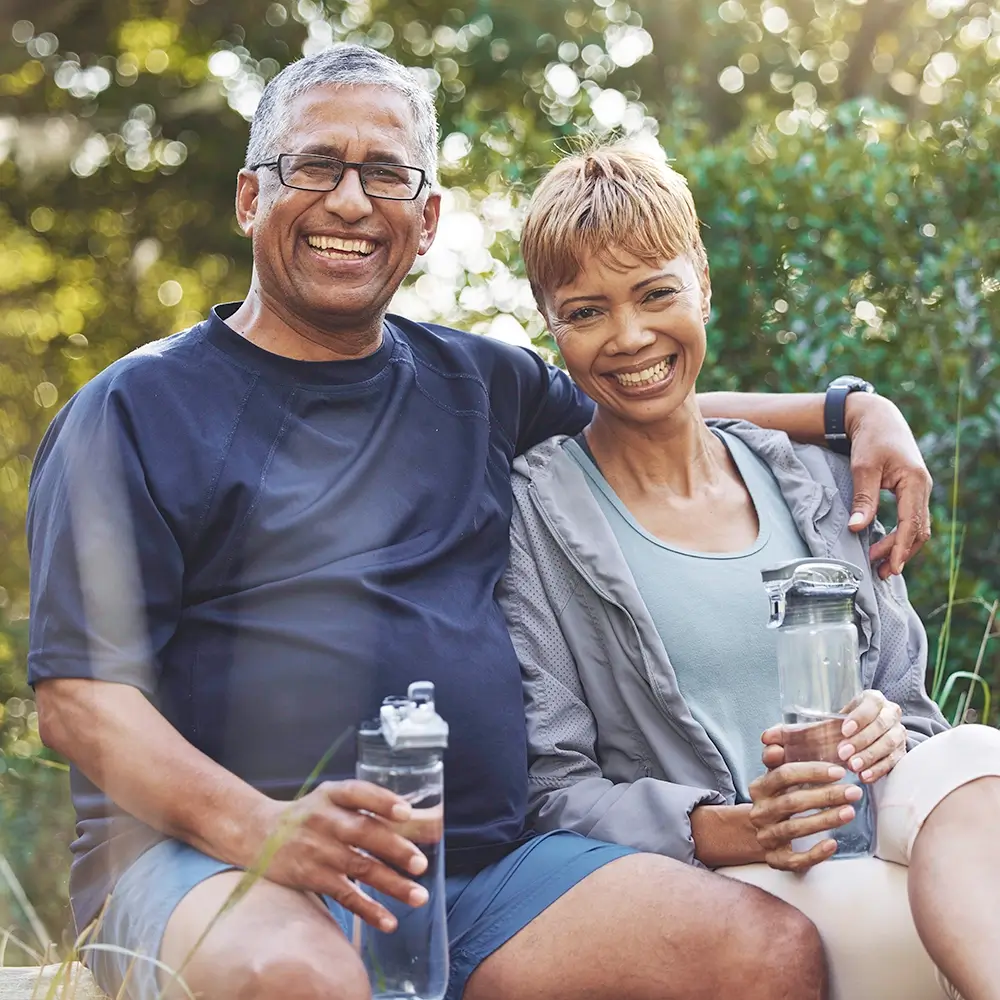 An older couple sits close together outdoors, smiling and holding water bottles, dressed in casual athletic wear, with greenery in the background.