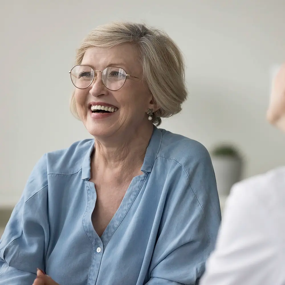 An older woman with short blonde hair and glasses smiles warmly. She is wearing a light blue blouse and sitting indoors, conversing with another person whose back is visible in the foreground.
