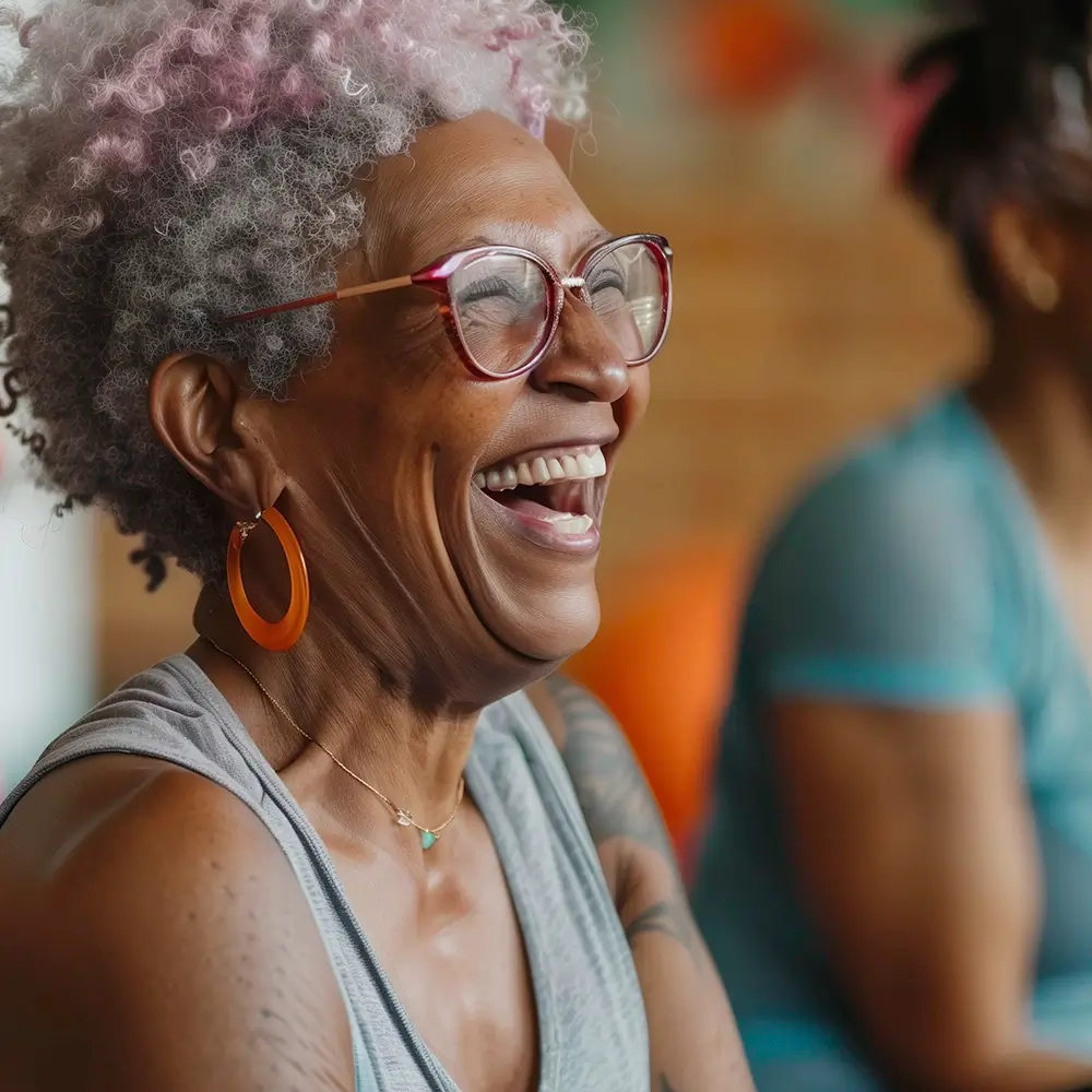 An older woman with curly gray and pink hair, wearing glasses, a sleeveless top, and large orange earrings, smiles and laughs. Another person is blurred in the background.
