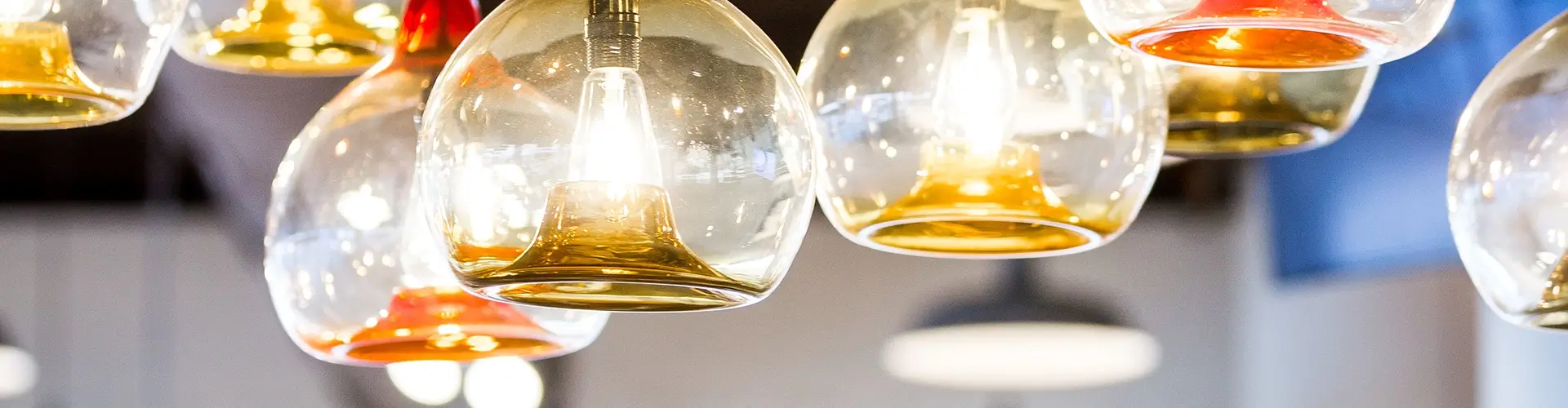 A close-up of several glass pendant lights with glowing bulbs inside, featuring clear and amber-tinted glass shades, hanging from the ceiling in a modern indoor setting.